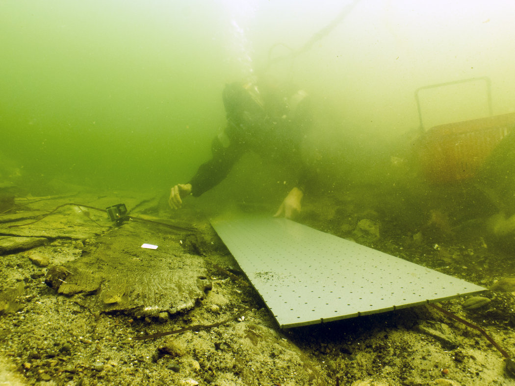 Marinarkæologen gør klar til at løfte de skrøbelige skibsdele ud af vandet på en tilpasset plade, som skibsdelen pakkes ind sammen med for at stabilisere og beskytte