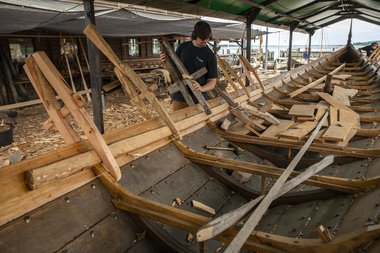 Bådebyggerlærling Magnus Ibsen Rishøj monterer en af de lange bjælkevægere. Under arbejdet holdes vægeren på plads med bordklemmer, mens placering og tilpasning kontrolleres langs skibets fulde længde, inden vægeren fastgøres med trænagler.