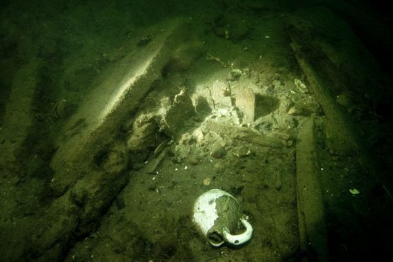 Glaseret lertøjskande, rester af spand eller pøs (bagerst i lysstrålen) og en del kraftigt tovværk (i bunden omkring kanden og tv. i billedet delvis under planken). Foto: Morten Johansen, Vikingeskibsmuseet