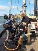 Tre af Vikingeskibsmuseets dykkende arkæologer. Det kræver hjælp at få monteret den tunge hjelm. Foto: Jørgen Dencker, Vikingeskibsmuseet. Tre af Vikingeskibsmuseets dykkende arkæologer. Det kræver hjælp at få monteret den tunge hjelm. Foto: Jørgen Dencker, Vikingeskibsmuseet.