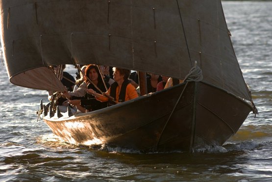 En aftensejlads på fjorden, hvor det gyldne aftenlys rammer bådens side, mens besætning putter sig under sejlets blafren