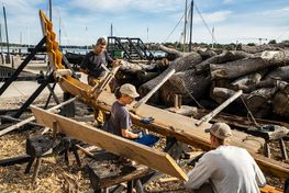 Boat builders work on the reconstruction of Skuldelev 5 outdoors in the sun. Copyright: The viking Ship museum