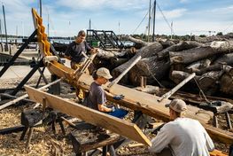 Bådebyggerne arbejder på Skuldelev 5's første bordgang i det smukke vejr.