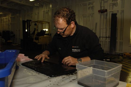 David Gregory, Nationalmuseets Bevaringsafdeling renser det sidste bundslam af låget til kobberkarret. Foto: Morten Johansen, Vikingeskibsmuseet. David Gregory, Nationalmuseets Bevaringsafdeling renser det sidste bundslam af låget til kobberkarret. Foto: Morten Johansen, Vikingeskibsmuseet.